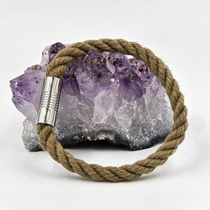 May include: A brown rope bracelet with a silver metal clasp. The bracelet is resting on a purple amethyst crystal.