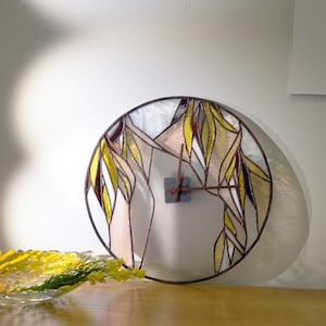 Puede incluir: Un reloj redondo de vidrieras con un marco de metal marrón. La esfera del reloj presenta un diseño de hojas amarillas y marrones. El reloj está montado en una pared blanca sobre un mueble de madera.