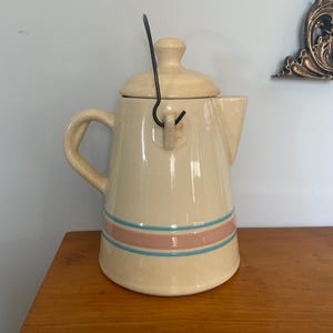 May include: Cream-colored ceramic teapot with a lid and handle. The teapot features a pink and blue stripe design around the middle. The handle is black. The teapot is sitting on a wooden surface.