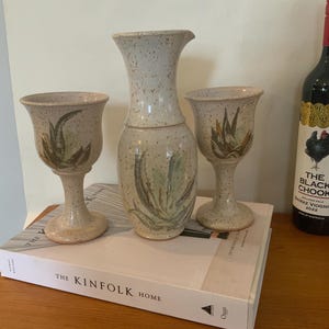 Peut inclure: Un pichet en céramique fait à la main et deux gobelets assortis avec une finition beige mouchetée. Chaque pièce est ornée d'un motif d'aloe vera vert et marron peint à la main. L'ensemble est présenté sur un livre intitulé "The Kinfolk Home".