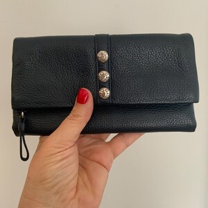 May include: Black leather wallet with a textured finish. The wallet has three silver decorative buttons and a wrist strap. The wallet is closed and appears to be empty.