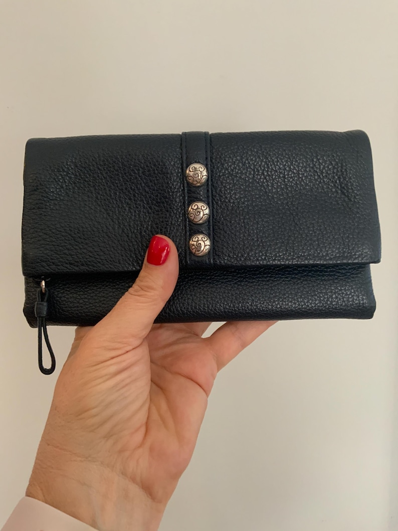 May include: Black leather wallet with a textured finish. The wallet has three silver decorative buttons and a wrist strap. The wallet is closed and appears to be empty.