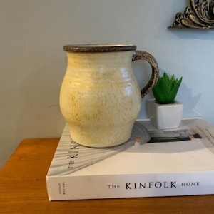 May include: A handcrafted ceramic mug with a pale yellow glaze, brown rim and handle. The mug rests on a white book titled "The Kinfolk Home", with a small green plant in a white pot beside it.