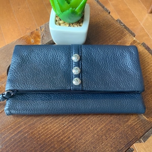 May include: A navy blue leather wallet with a textured finish. The wallet features a flap closure fastened with three silver decorative buttons. A small, green plant in a white square pot is on a wooden surface.
