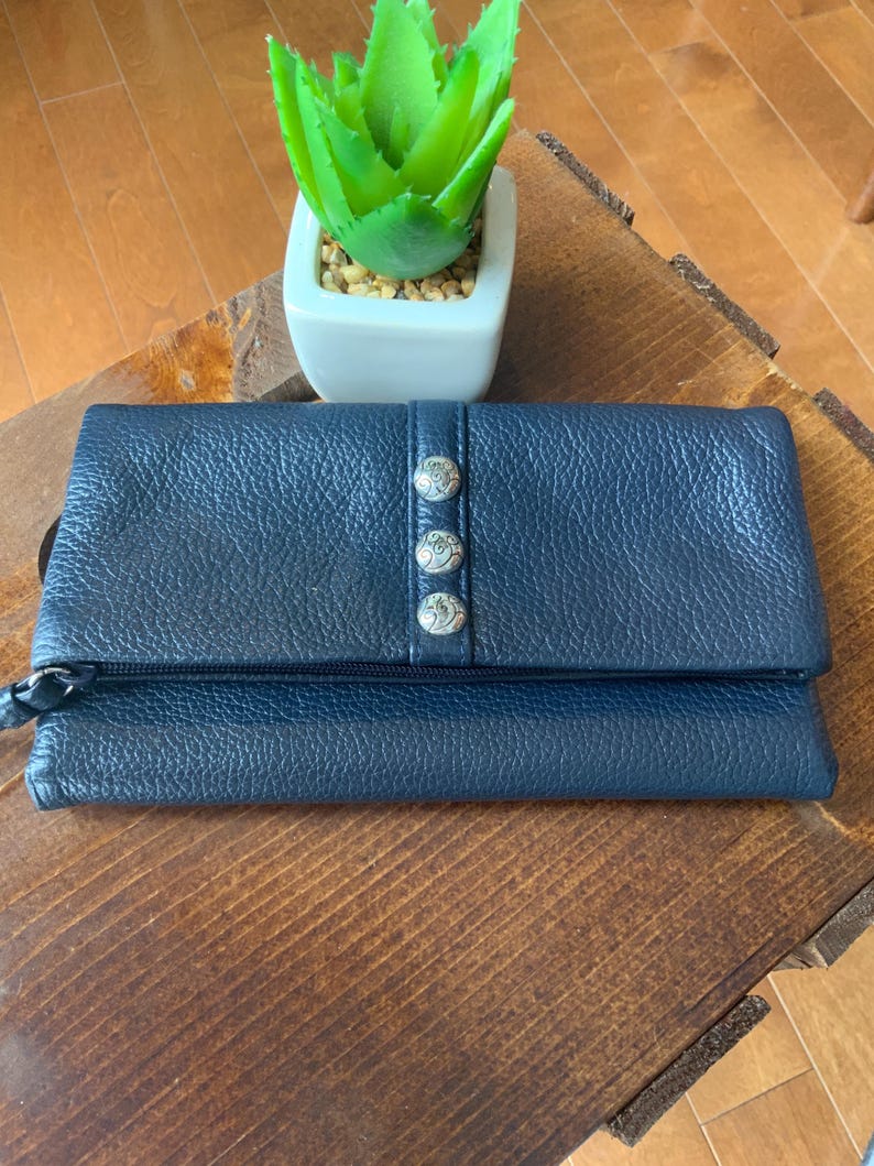 May include: A navy blue leather wallet with a textured finish. The wallet features a flap closure fastened with three silver decorative buttons. A small, green plant in a white square pot is on a wooden surface.
