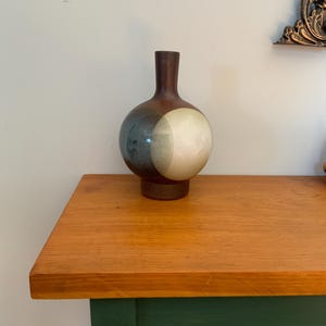 May include: A spherical ceramic vase with a narrow neck and a flat base. The body features a dark brown finish at the top and bottom, with two overlapping circular patterns in shades of charcoal grey and off-white on the centre of the vase.