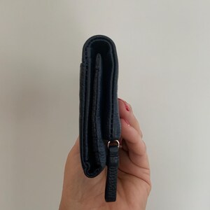 May include: A navy blue leather wallet with a textured finish. The wallet has a small loop and a leather pull tab. The wallet is closed and held vertically.