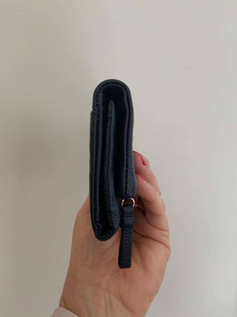 May include: A navy blue leather wallet with a textured finish. The wallet has a small loop and a leather pull tab. The wallet is closed and held vertically.