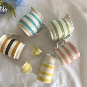 May include: A collection of ceramic pitchers and small cups with a cream base and colorful stripes. The pitchers have a handle and spout. Stripes include blue, green, pink, brown, and yellow. The cups and a tall container also have yellow stripes.