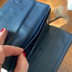 May include: A navy blue leather wallet, open to show internal pockets and card slots. The wallet has a textured surface and a snap closure. The interior lining is a lighter shade of blue.