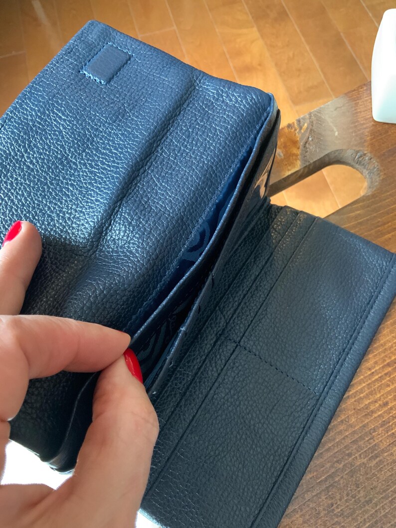 May include: A navy blue leather wallet, open to show internal pockets and card slots. The wallet has a textured surface and a snap closure. The interior lining is a lighter shade of blue.