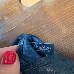 May include: Close-up of a dark blue leather glove with the brand name "Brighton" on a black tag. The glove has a textured surface and visible stitching. The background is a wooden surface.