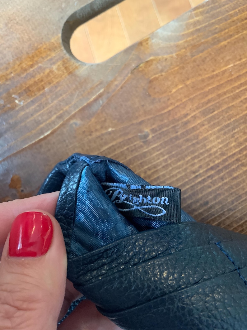 May include: Close-up of a dark blue leather glove with the brand name "Brighton" on a black tag. The glove has a textured surface and visible stitching. The background is a wooden surface.