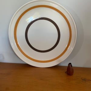 May include: A white ceramic plate with a brown and orange concentric circle design. A small, brown, pear-shaped object sits to the side of the plate. The plate has a simple, mid-century modern aesthetic.
