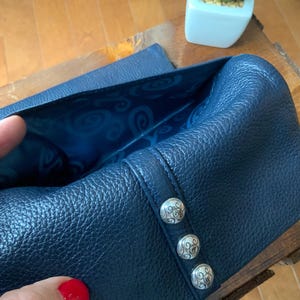 May include: A navy blue leather wallet with silver decorative buttons. The interior lining is a lighter blue with a swirling pattern. The wallet is open, revealing the inside. A small succulent plant in a white square pot is in the background.
