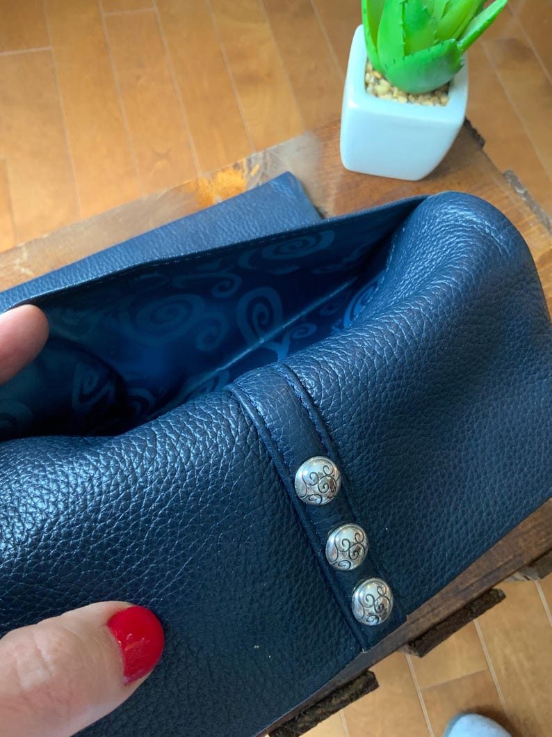 May include: A navy blue leather wallet with silver decorative buttons. The interior lining is a lighter blue with a swirling pattern. The wallet is open, revealing the inside. A small succulent plant in a white square pot is in the background.