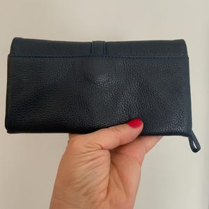 May include: A navy blue leather wallet with a textured finish. The rectangular wallet has a loop on the side. The wallet is held in a hand.