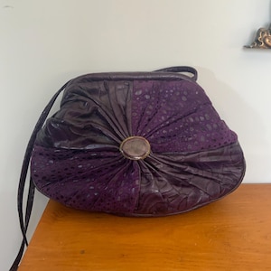 May include: A vintage purple handbag with a distinctive design. The bag has a central section of textured, patterned fabric, surrounded by pleated, dark purple leather. A circular clasp is in the centre, and a thin strap extends from the top.