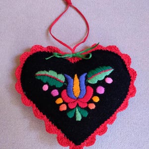 Peut inclure: Un coussin en forme de cœur en feutre noir avec une bordure en crochet rouge. Le coussin est brodé d'un motif floral coloré dans les tons de vert, bleu, orange, rose et rouge.