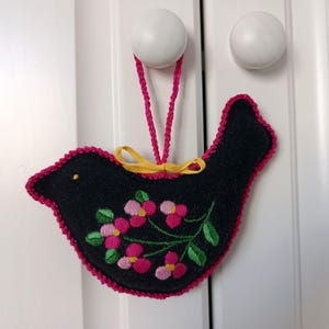 Könnte beinhalten: Schwarzer Vogelanhänger aus Filz mit rosa gestickten Blumen und einer gelben Schleife. Der Vogel hat einen rosa gestickten Rand und eine rosa Kordel zum Aufhängen. Der Anhänger hängt an einer weißen Tür.