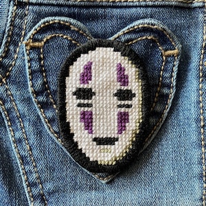 Op de afbeelding: Een witte en zwarte geborduurde patch van No Face uit de film Spirited Away, genaaid op een blauwe denim stof.
