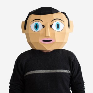 May include: A papercraft mask of a person's face with large blue eyes and a pink mouth. The mask is made of brown and white paper and has a black hair line.
