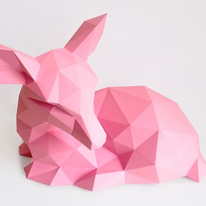 May include: A pink origami deer sculpture, made of paper, with a geometric design. The deer is lying down with its head turned to the side.