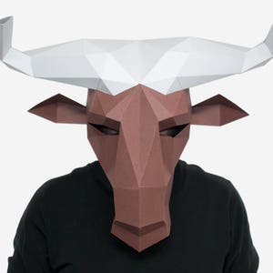 Puede incluir: Una máscara de papel de cabeza de toro marrón y blanca. La máscara está hecha de papel doblado y tiene un diseño realista.