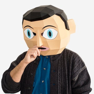 以下が含まれることがあります： 驚いた表情をした紙粘土のマスクをつけた人。マスクはベージュで、黒い髪、青い目、ピンクの口をしています。その人は、青いボタンダウンシャツの上に濃いグレーのセーターを着ています。