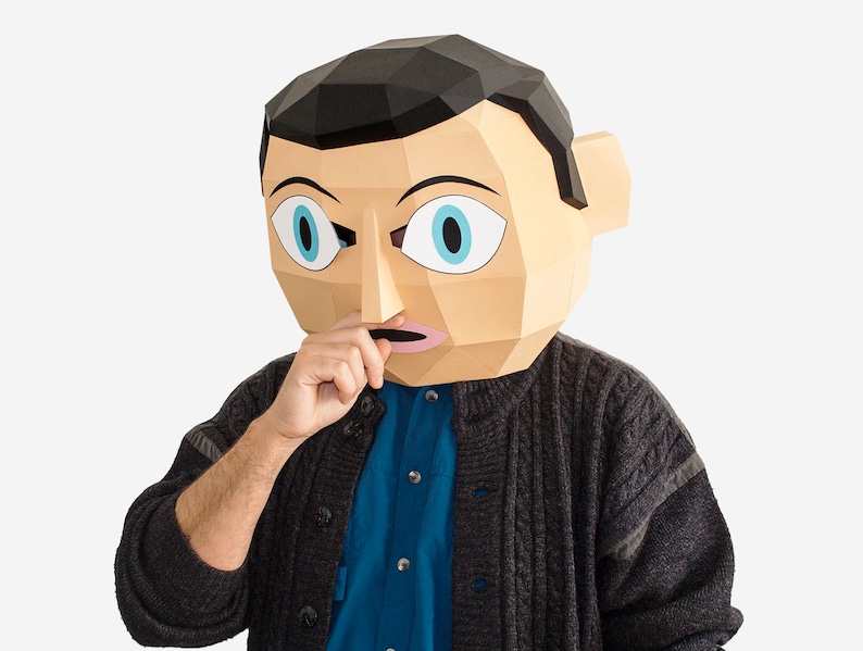 May include: A person wearing a paper mache mask with a surprised expression. The mask is beige with black hair, blue eyes, and a pink mouth. The person is wearing a dark gray sweater over a blue button-down shirt.