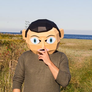 May include: A person wearing a paper mache mask of a face with blue eyes and a pink nose. The mask is covering the person's face and they are holding their hand over their mouth. The person is standing in front of a grassy area with a body of water in the background.