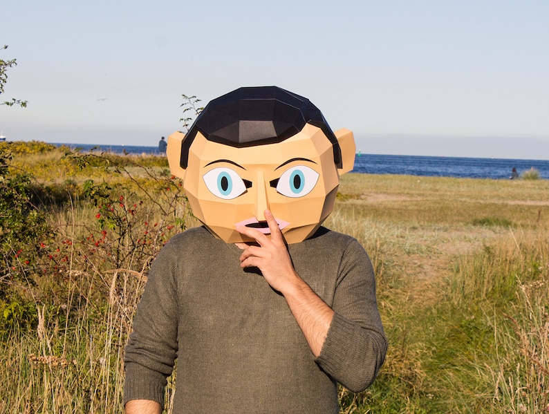 May include: A person wearing a paper mache mask of a face with blue eyes and a pink nose. The mask is covering the person's face and they are holding their hand over their mouth. The person is standing in front of a grassy area with a body of water in the background.