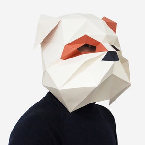 May include: A white paper mask shaped like a bulldog's head with orange ears and a black nose. The mask is folded into a three-dimensional shape.