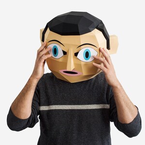 May include: A person wearing a paper mache mask with large blue eyes and a surprised expression. The mask is brown and beige with black hair. The person is wearing a gray sweater with a white stripe.
