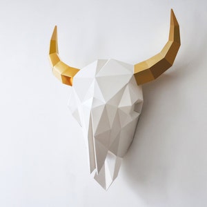 K&ouml;nnte beinhalten: Ein wei&szlig;er Papiermach&eacute;-Stierkopf mit goldenen H&ouml;rnern. Der Sch&auml;del besteht aus geometrischen Formen und ist ein 3D-Wanddekor.