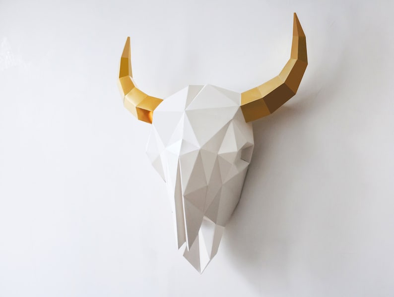 K&ouml;nnte beinhalten: Ein wei&szlig;er Papiermach&eacute;-Stierkopf mit goldenen H&ouml;rnern. Der Sch&auml;del besteht aus geometrischen Formen und ist ein 3D-Wanddekor.