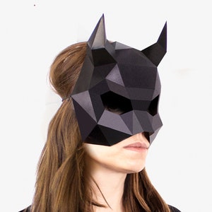 Könnte beinhalten: Eine schwarze Papiermaske mit geometrischem Design und zwei Hörnern. Die Maske wird von einer Person mit langen braunen Haaren getragen.