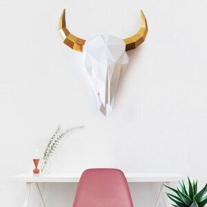 K&ouml;nnte beinhalten: Ein wei&szlig;er Papier-Stierkopf mit goldenen H&ouml;rnern, der an einer wei&szlig;en Wand &uuml;ber einem wei&szlig;en Schreibtisch mit einem roten Stuhl und einer gr&uuml;nen Pflanze montiert ist.
