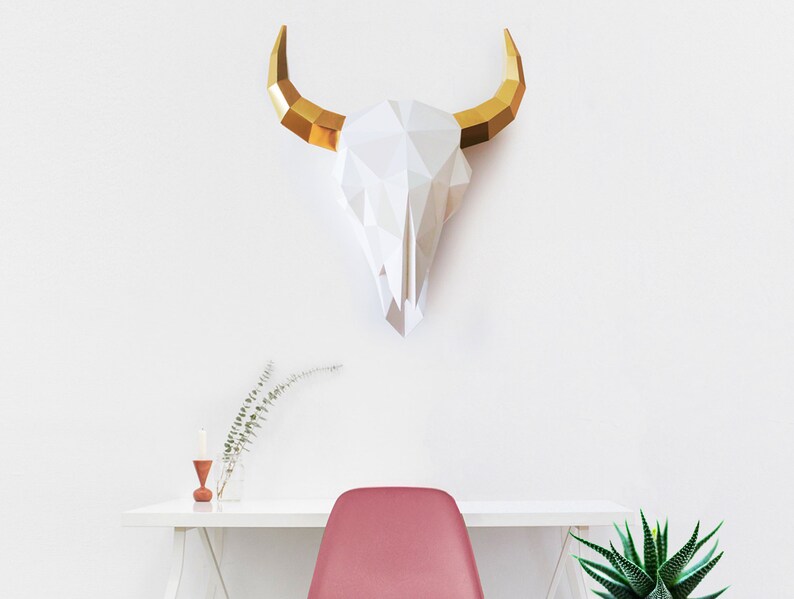 K&ouml;nnte beinhalten: Ein wei&szlig;er Papier-Stierkopf mit goldenen H&ouml;rnern, der an einer wei&szlig;en Wand &uuml;ber einem wei&szlig;en Schreibtisch mit einem roten Stuhl und einer gr&uuml;nen Pflanze montiert ist.