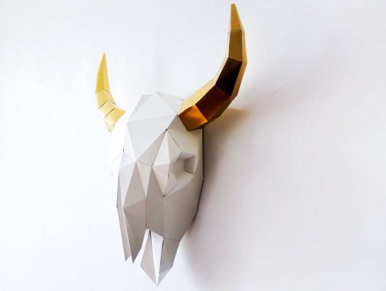 K&ouml;nnte beinhalten: Ein wei&szlig;er Papier-Stierkopf mit goldenen H&ouml;rnern. Der Sch&auml;del besteht aus gefaltetem Papier und hat ein geometrisches Design.