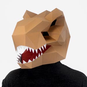 Puede incluir: Una máscara de dinosaurio de papel maché marrón con una boca roja y dientes blancos. La máscara la lleva puesta una persona con una camiseta negra.