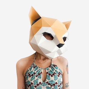 May include: A person wearing a paper mache fox mask with a geometric design. The mask is white and orange with black accents. The person is wearing a blue and green patterned halter top.