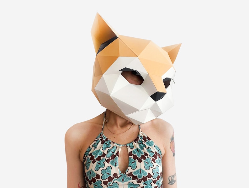 May include: A person wearing a paper mache fox mask with a geometric design. The mask is white and orange with black accents. The person is wearing a blue and green patterned halter top.