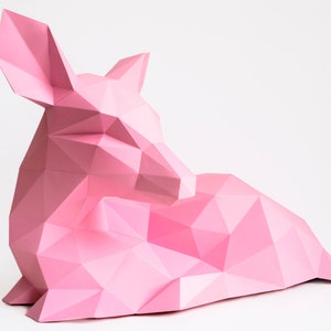 May include: A pink origami deer sculpture with a geometric design. The deer is lying down with its head resting on its front legs.