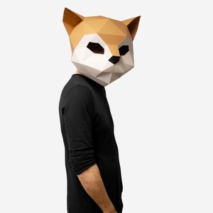 May include: A person wearing a brown and white paper mache fox mask. The mask has a geometric design and is covering the person's face. The person is wearing a black long-sleeved shirt.