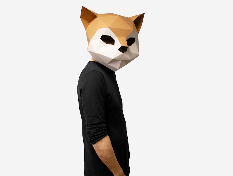 May include: A person wearing a brown and white paper mache fox mask. The mask has a geometric design and is covering the person's face. The person is wearing a black long-sleeved shirt.