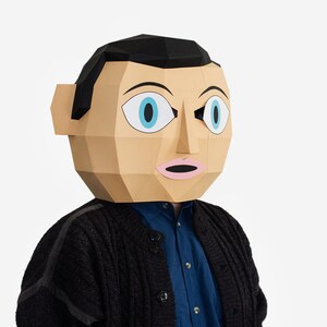 May include: A person wearing a paper mache mask with a black hair, blue eyes, and a pink mouth. The mask is in the shape of a human face. The person is wearing a black sweater over a blue shirt.