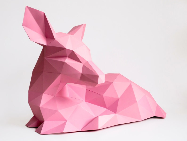 May include: A pink papercraft deer sculpture in a lying down position. The deer is made of folded paper and has a geometric design.