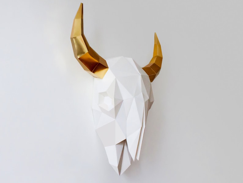 K&ouml;nnte beinhalten: Ein wei&szlig;er Papier-Stierkopf mit goldenen H&ouml;rnern. Der Sch&auml;del besteht aus gefaltetem Papier und hat ein geometrisches Design.