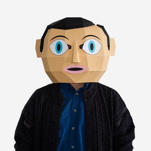 May include: A person wearing a paper mache mask with a surprised expression. The mask is brown with blue eyes and pink lips. The person is wearing a black sweater over a blue shirt.